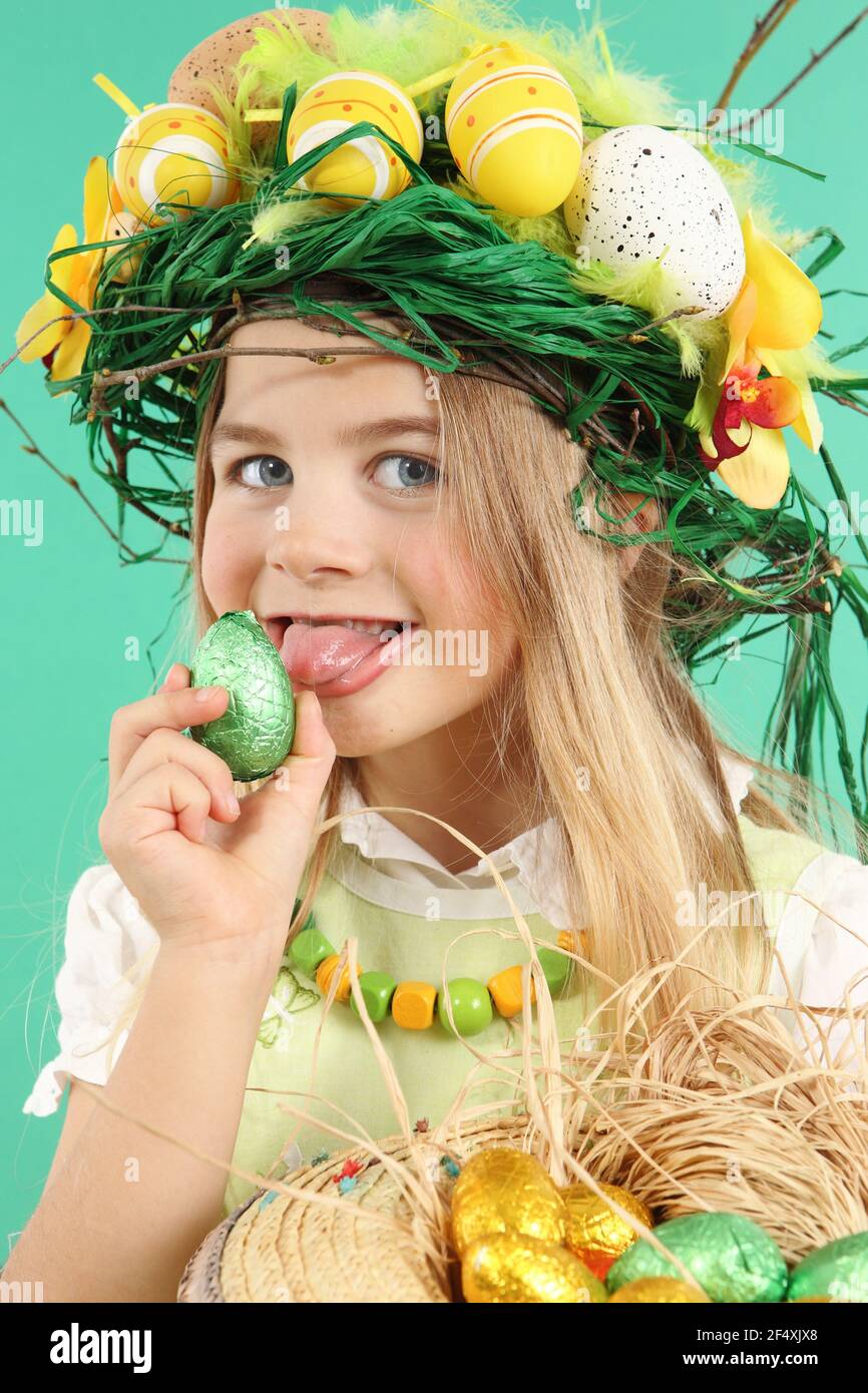 Portrait of a charming young girl wearing a festive hairstyle made with yellow Easter eggs, spring flowers and bird feathers. The child licks an easte - Stock Image