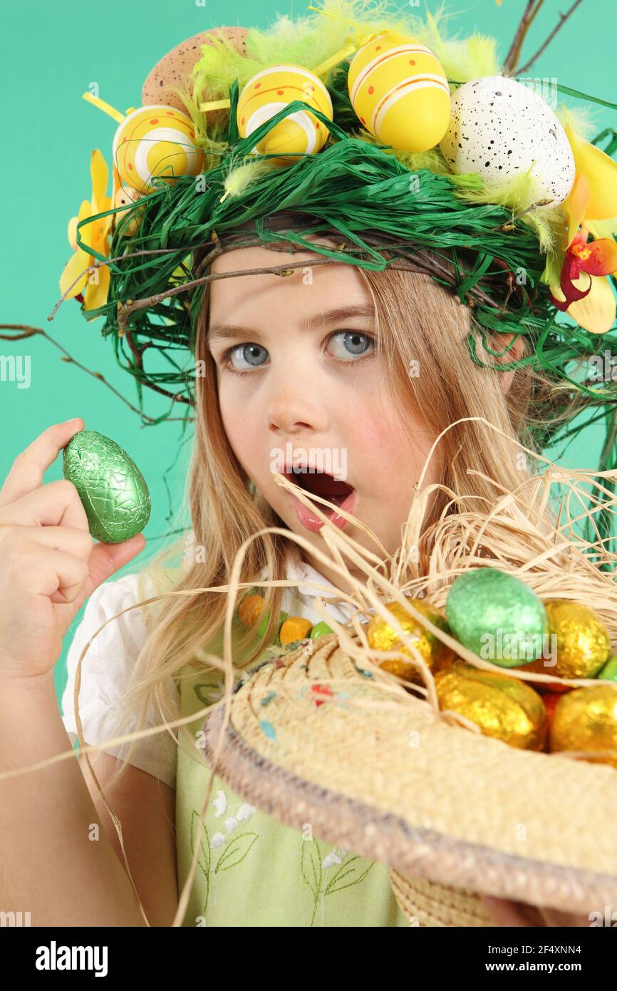 Young girl with Easter eggs basket egg is holding an egg in her hand looking at the camera. The girl is dressed for the Easter holidays with a straw b - Stock Image