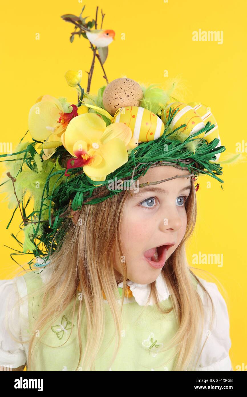 Easter concept on yellow background: Portrait of 7 year old child dressed with bird's nest hairstyle, made of spring flowers Easter eggs and feathers. - Stock Image