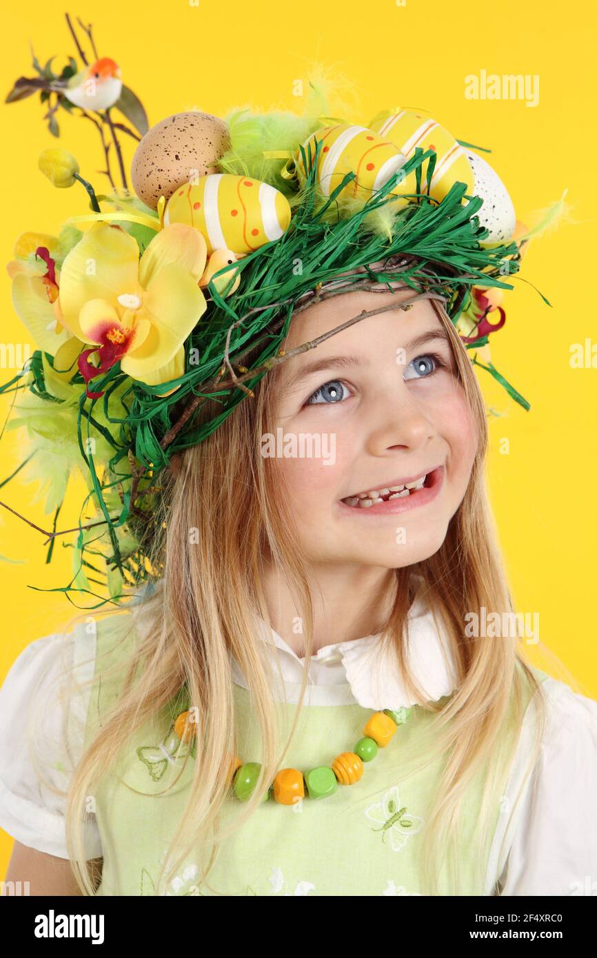 Happy girl having fun Easter egg hunt, wearing straw nest hairstyle made of spring flowers, Easter eggs and feathers. The child looks to the side smil - Stock Image