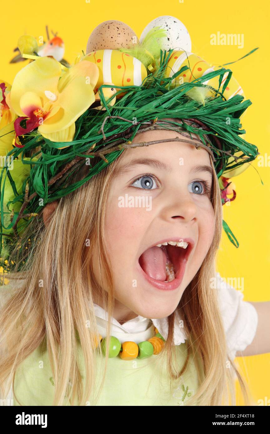 Young happy girl wearing straw headdress made of spring flowers, Easter eggs and feathers. The child with the big eyes is surprised by happiness. Art - Stock Image