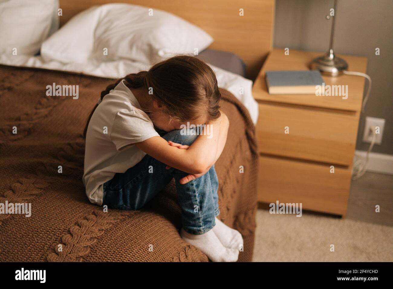 Top view of upset little girl hugging knee, sobbing with head bowed and crying sitting alone on bed in bedroom. Stock Photo