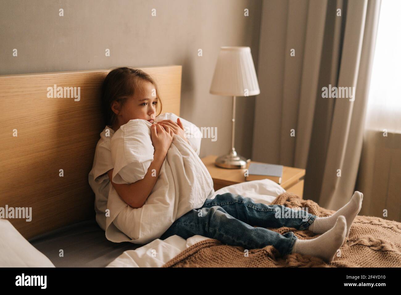 Upset little girl crying sitting alone in fetal position hiding face and rocking on bed hugging big pillow. Stock Photo