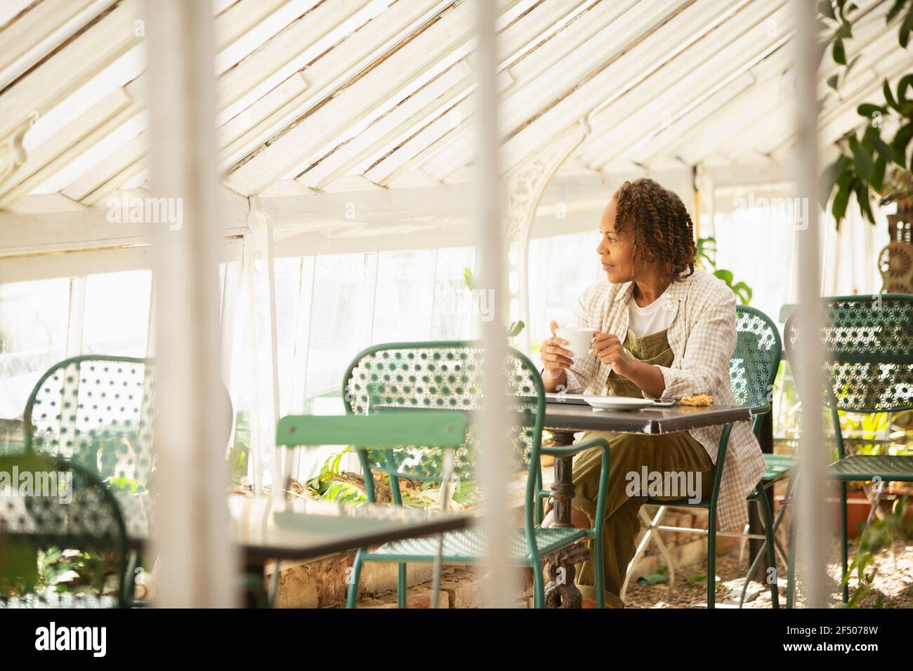 Female garden shop owner drinking tea at table in greenhouse Stock Photo