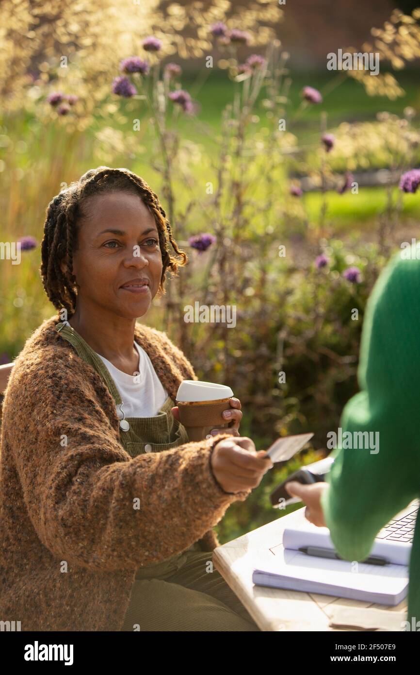 Woman with coffee paying waitress with smart card on sunny cafe patio Stock Photo