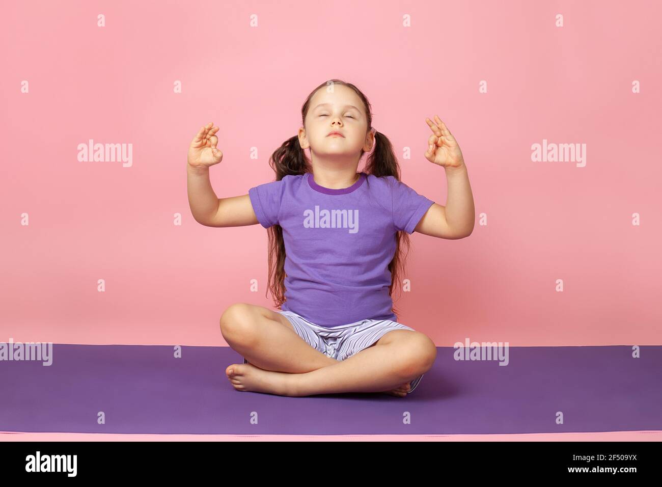 a girl with her eyes closed does yoga, sits in the lotus position or prays on a purple mat, isolated on a pink background Stock Photo