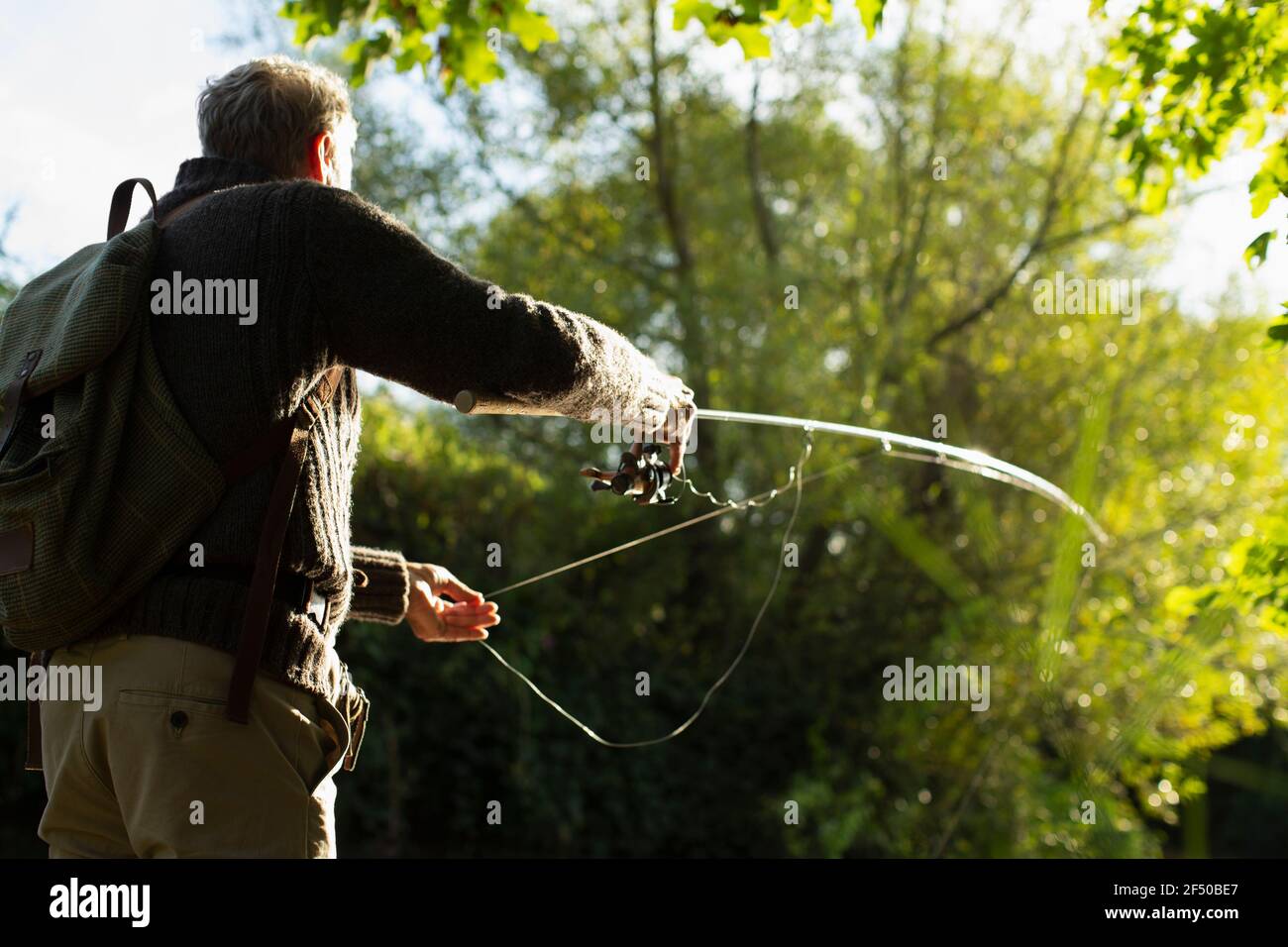 Man with backpack casting fly fishing pole below sunny trees Stock Photo -  Alamy