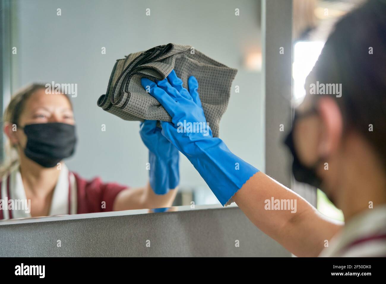 Female hotel maid in face mask and glove cleaning bathroom mirror Stock Photo