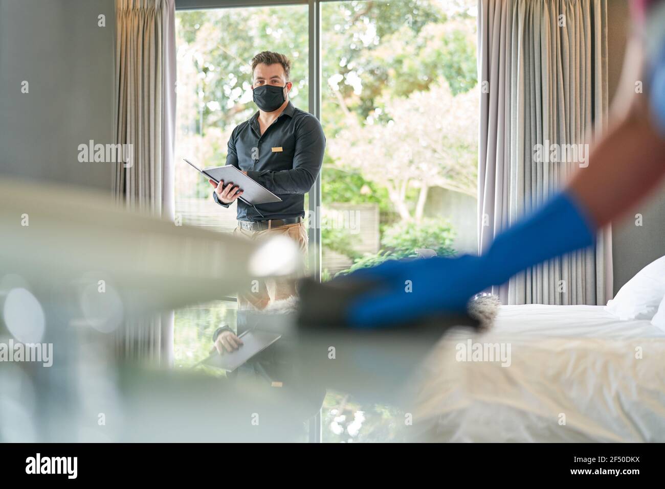 Hotel manager in face mask inspecting room and watching maid clean Stock Photo