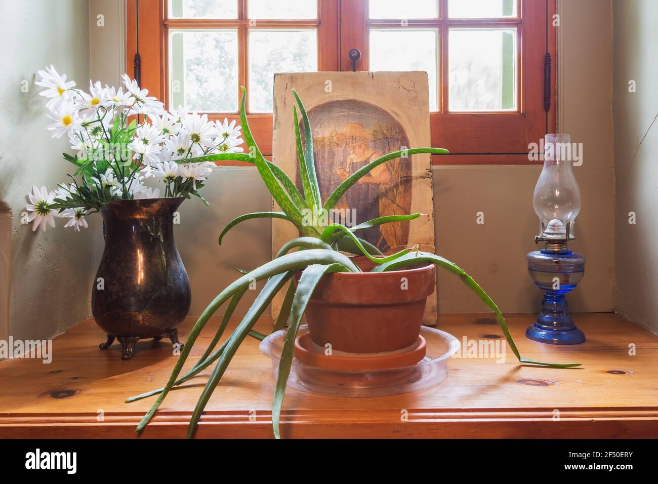White flowers in vintage vase, succulent plant in terracotta planter, framed painting of boy fishing, old blue oil lamp on windowsill inside old house Stock Photo