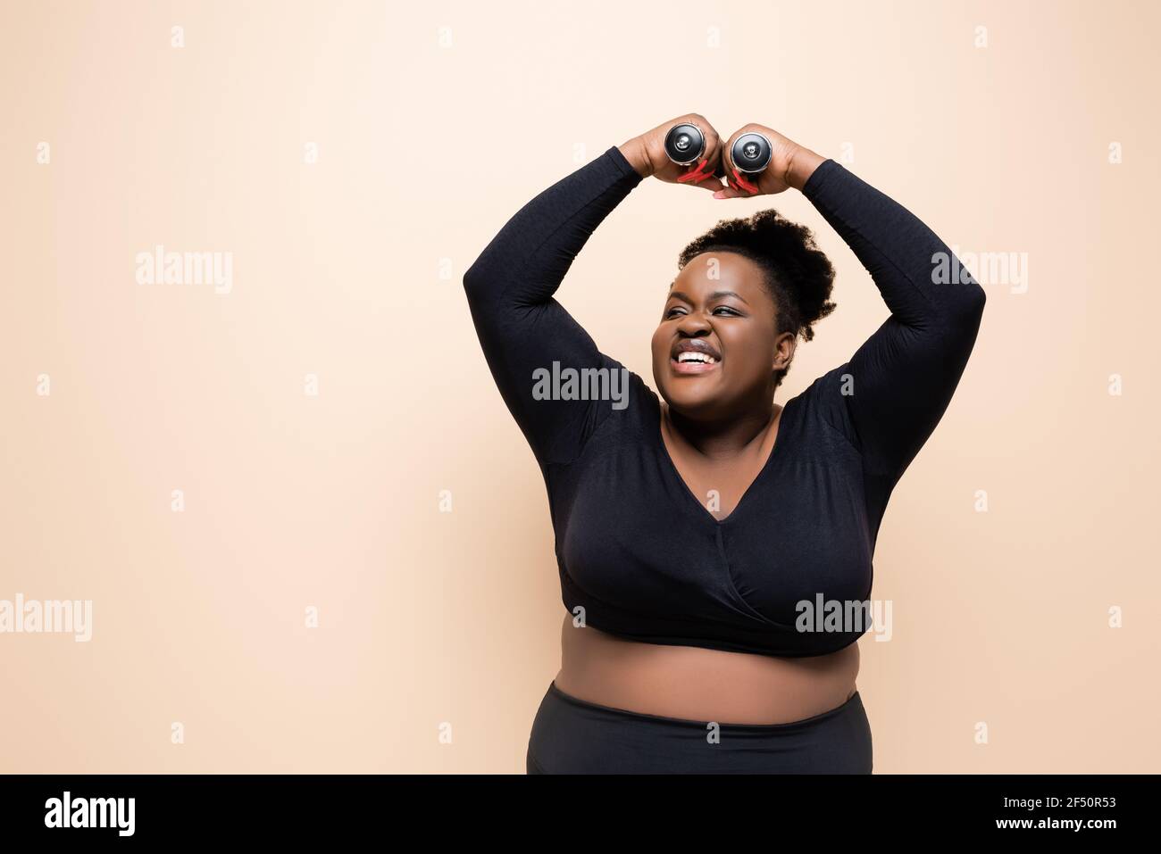 happy african american plus size woman in sportswear holding dumbbells above head isolated on beige Stock Photo