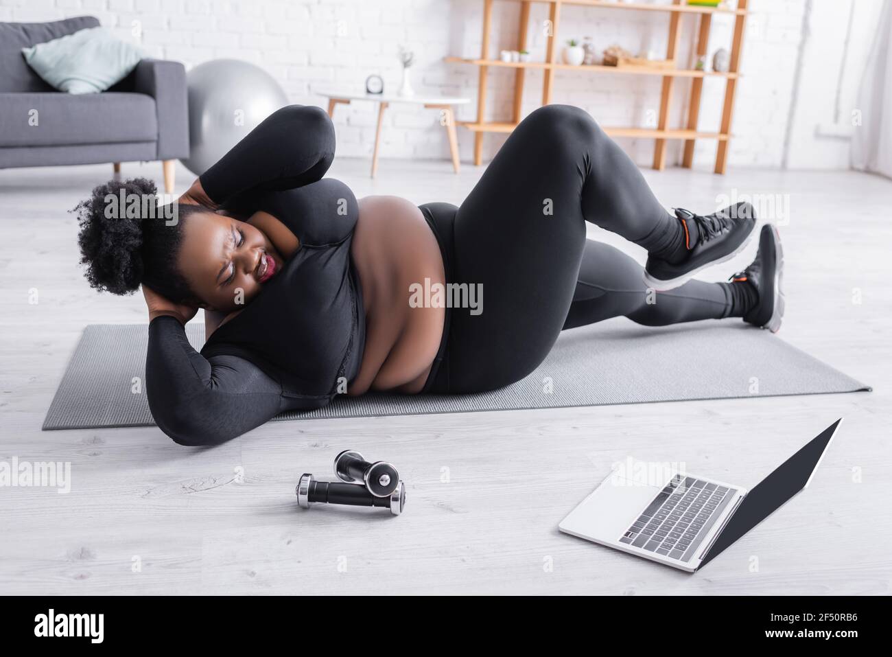 happy african american plus size woman in sportswear working out on fitness mat and looking at laptop Stock Photo