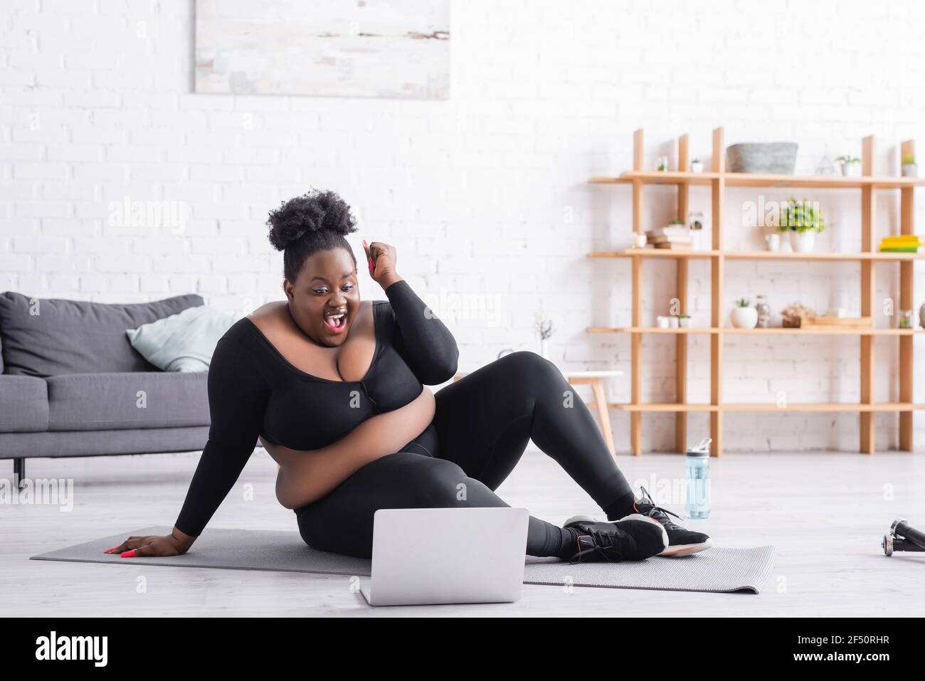 excited african american plus size woman in sportswear sitting on fitness mat and looking at laptop in living room Stock Photo