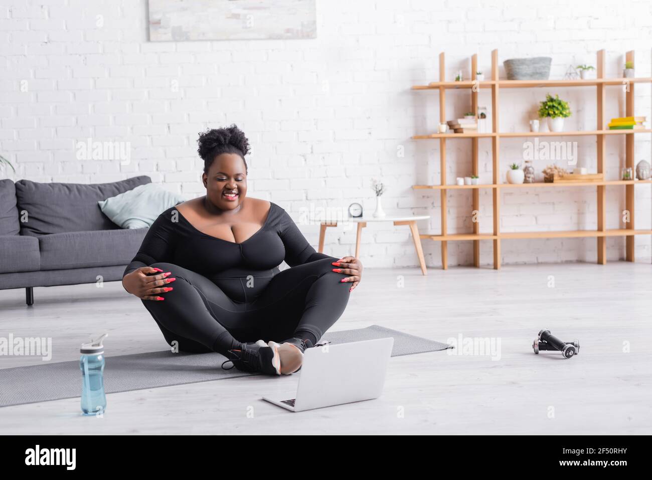 happy african american plus size woman in sportswear exercising near laptop in living room Stock Photo