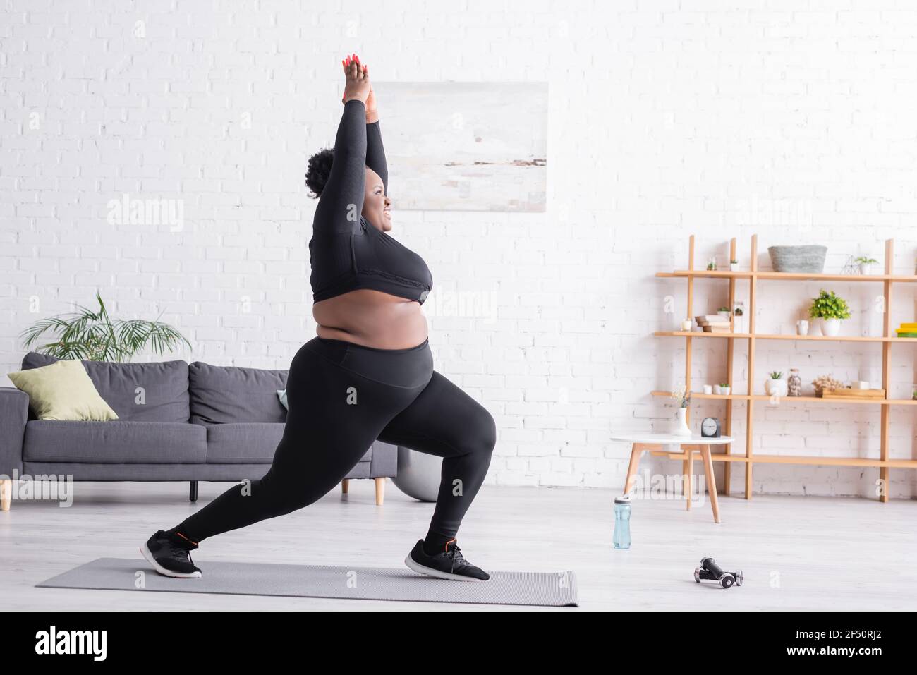 full length of cheerful african american plus size woman in sportswear standing in yoga pose in living room Stock Photo