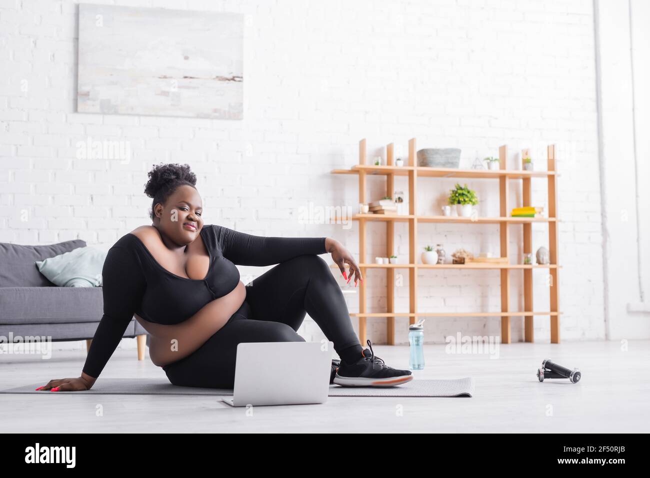 smiling african american plus size woman watching online training on laptop while sitting on fitness mat Stock Photo