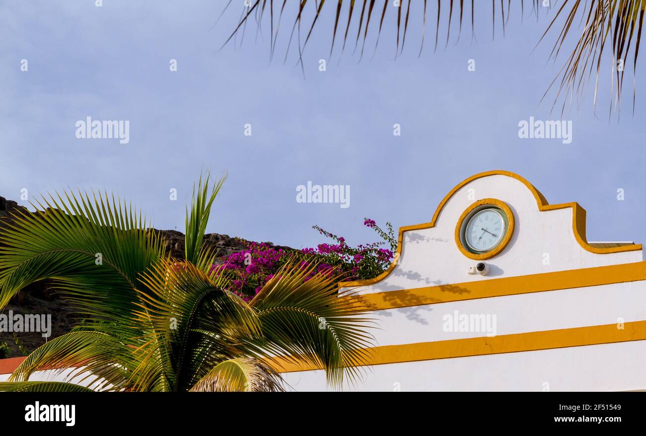 Detail of a clock in a white wall with yellow details by a palm tree on a sunny day Stock Photo