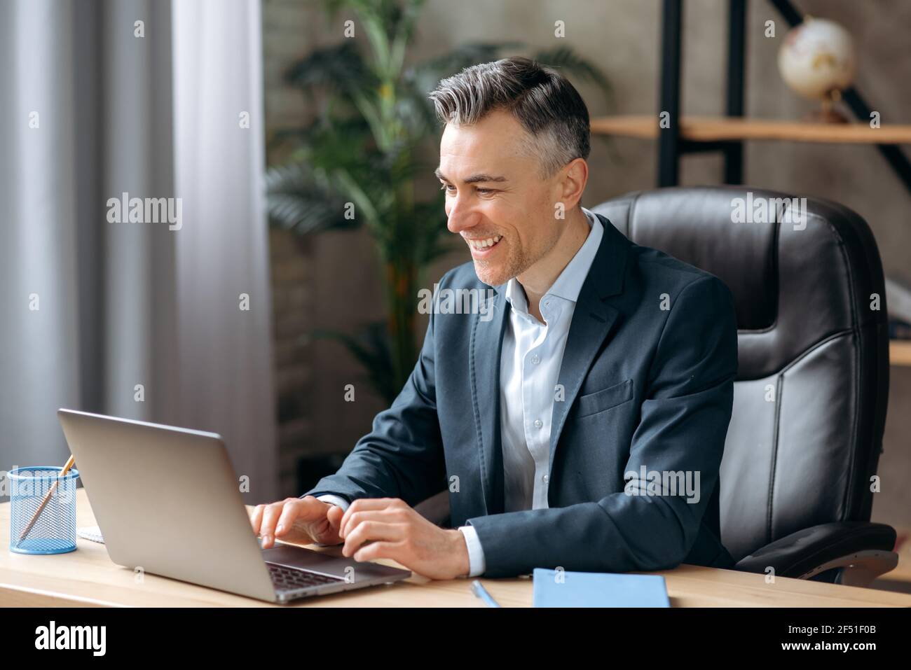 Influential successful Caucasian adult businessman or manager in a formal suit uses a laptop, communicates via video conferencing, online meetings, sits office, types on a keyboard, smiles friendly Stock Photo