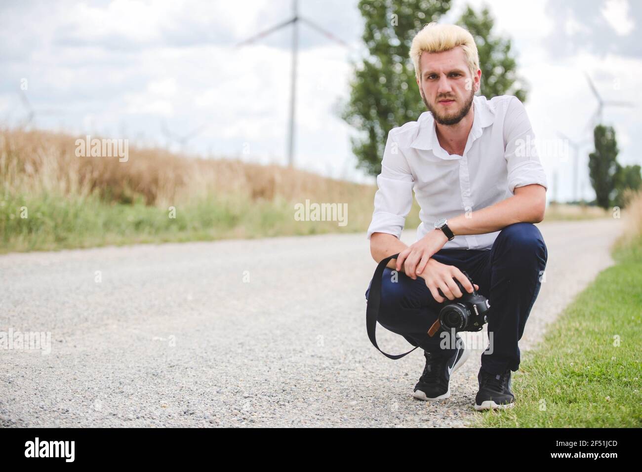 Young man squatting down at the edge of a road with different colored hair and beard with a camera Stock Photo