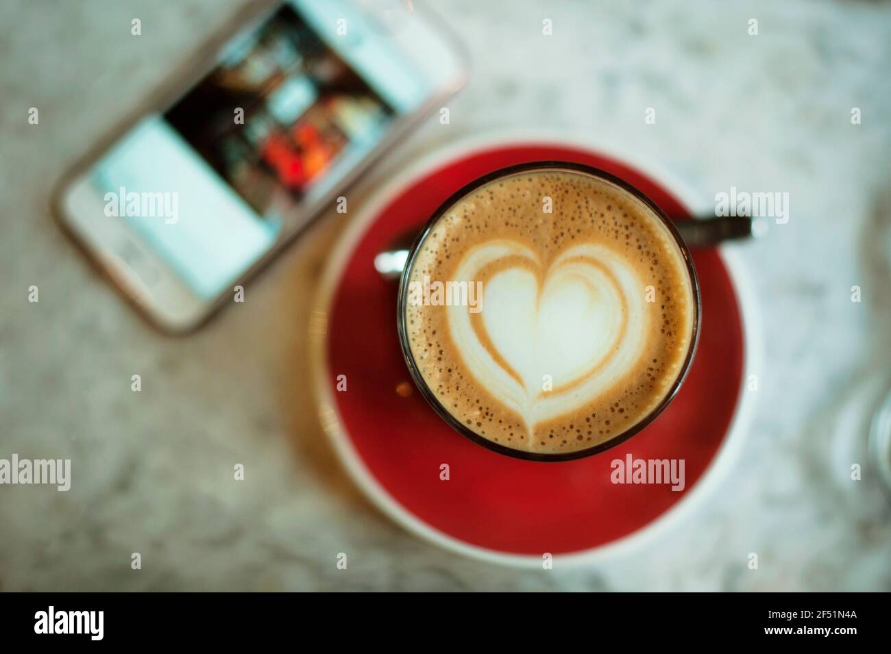 Close-up of coffee cup from above with a heart shape in froth and a smartphone on marble tabletop. Love coffee / coffee break concept Stock Photo