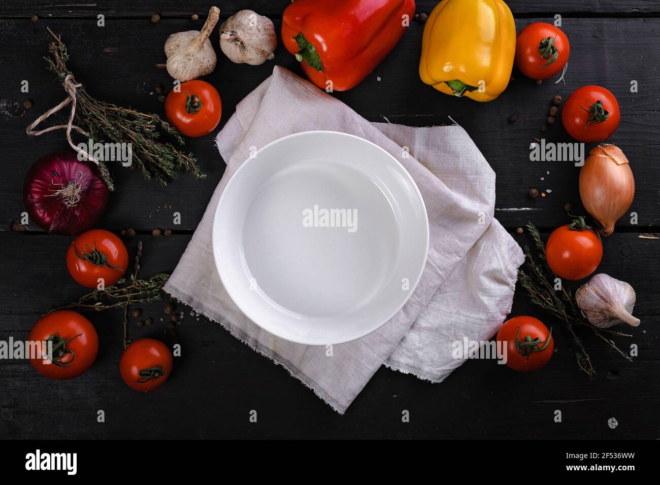 White clean plate on a napkin, bell peppers, tomatoes, garlic and onions are laying on a dark wooden table. Contrast bright colored flatlay. Top view. Stock Photo