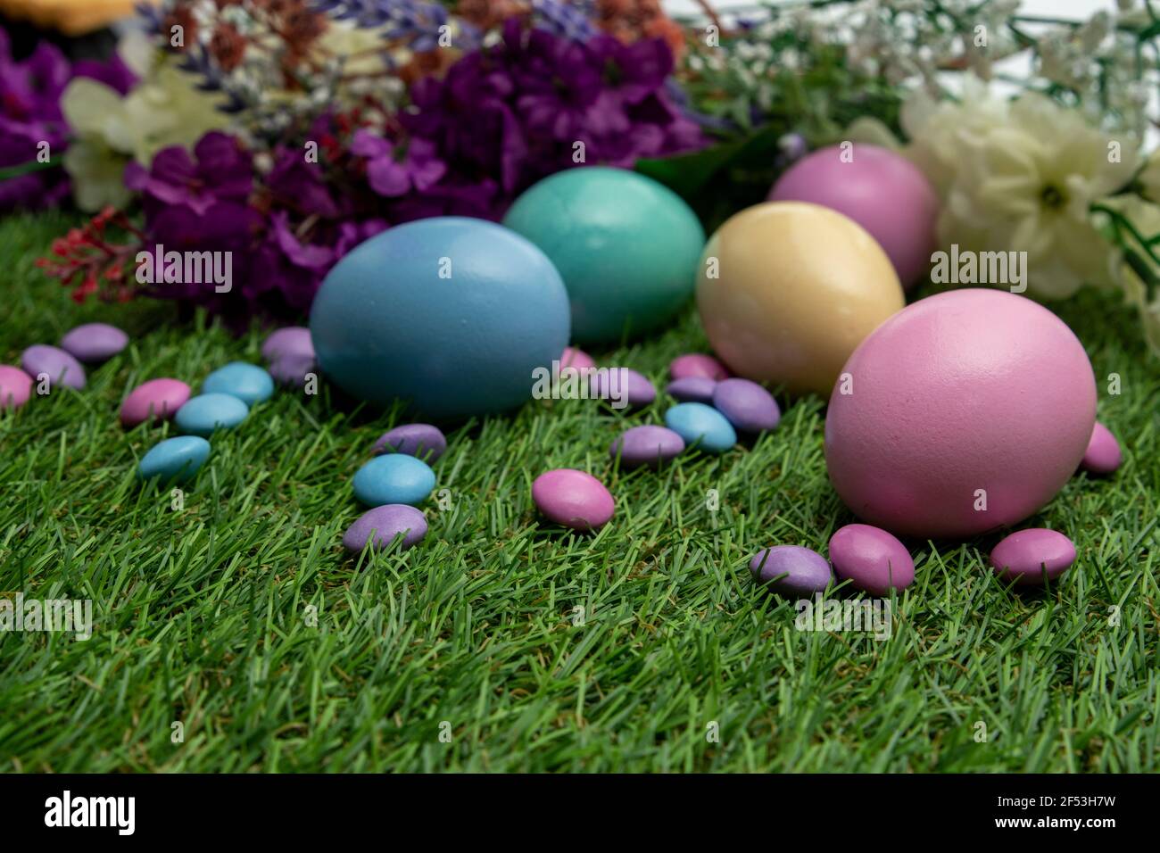 Pastel colored Easter eggs and pastel colored chocolate candy on green grass , against colored flowers background. Stock Photo