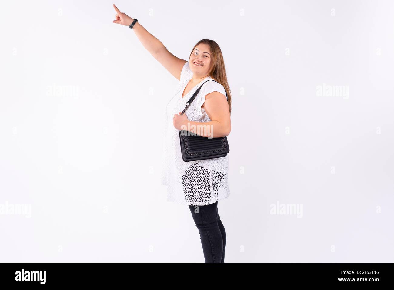 A plus size caucasian woman looks into the camera with a smile and points her index finger to the top. White studio background. Full-length photo with Stock Photo