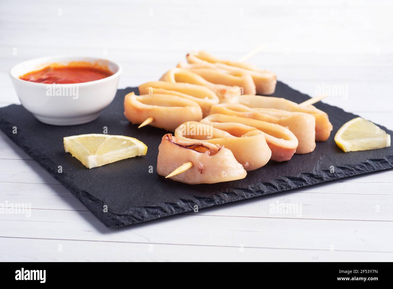 Fried squid rings on a skewer with tomato sauce and lemon. Black slate stand, white background copy space Stock Photo