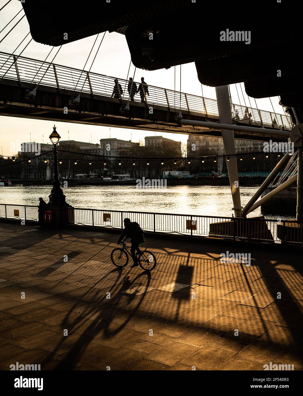 An evening scene on the south bank of the River Thames, London. Evening light creates shadows of people enjoying the sun. A cyclist and pedestrians Stock Photo