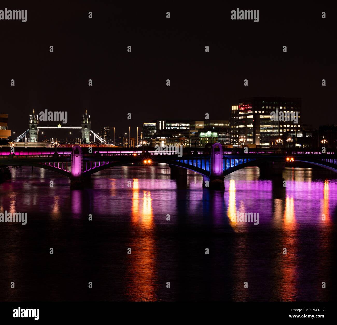 The view along the Thames to London Bridge illuminated at night with orange and purple lights reflected in the river Stock Photo