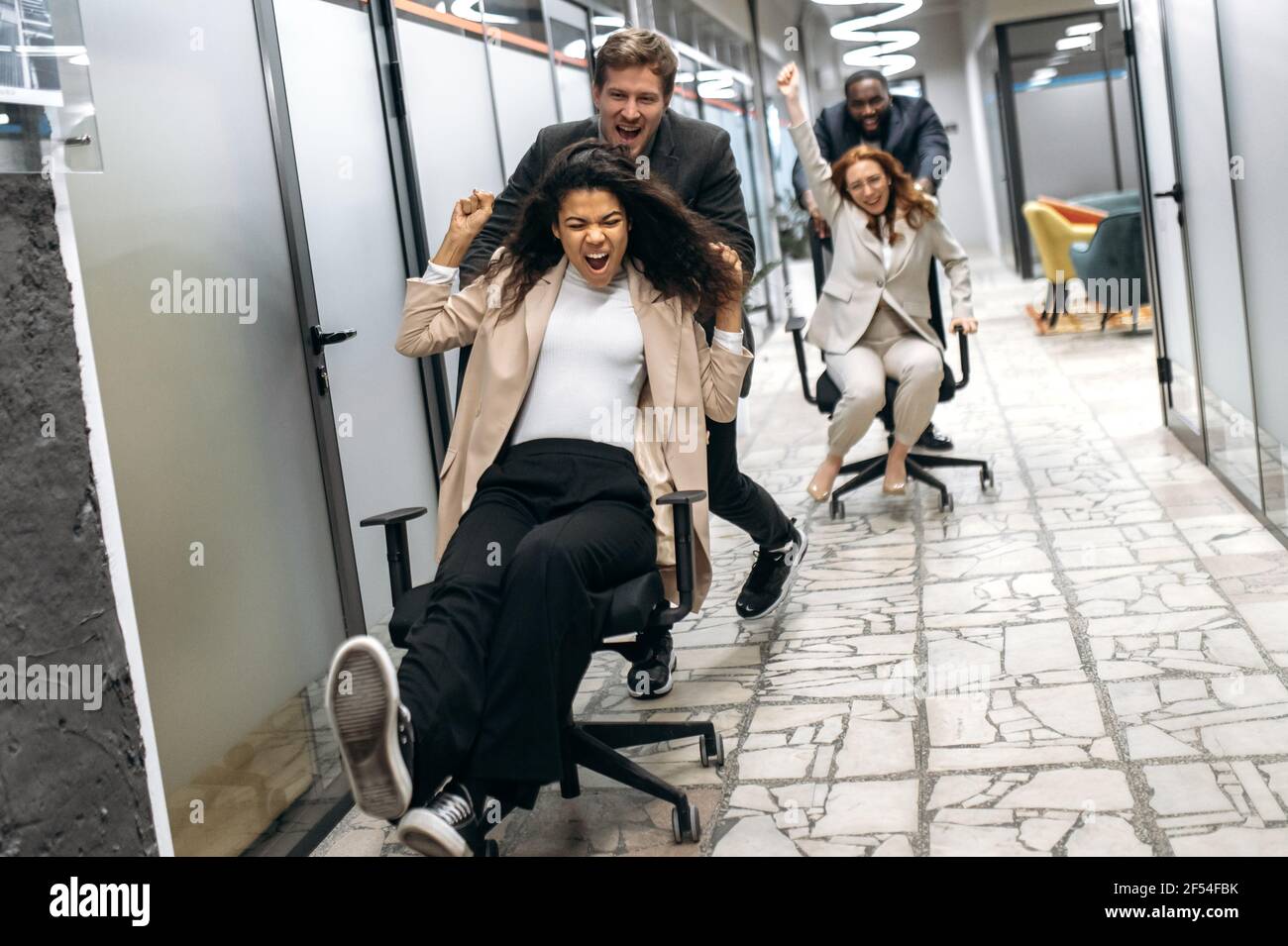 Emotional multiracial male and female employees having fun at the workplace. Young adult business people in formal wear making race on chairs in modern office while break Stock Photo