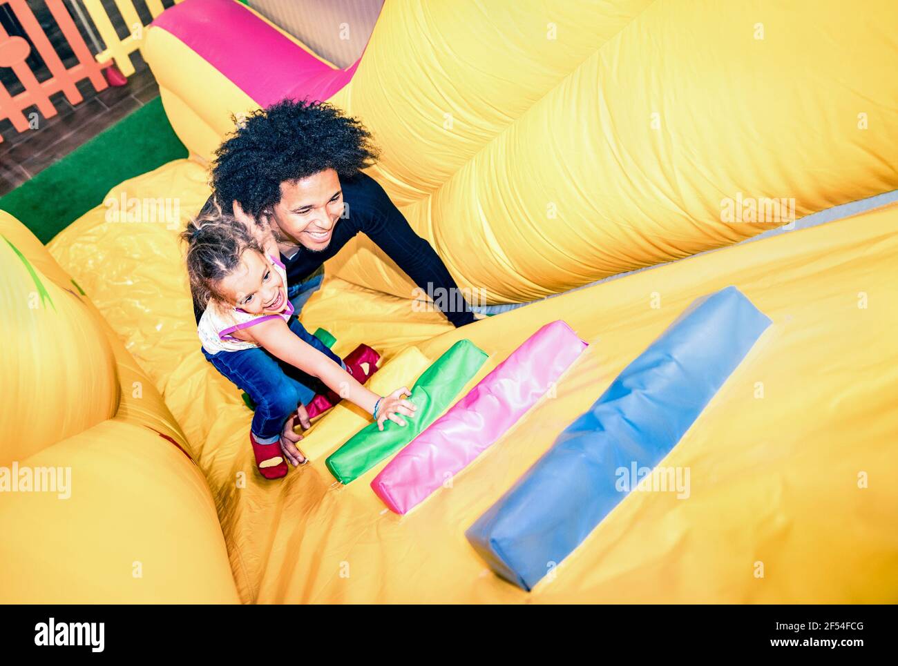Latin american dad playing with mixed race daughter on inflatable slide at kindergarten playroom - Family concept Stock Photo