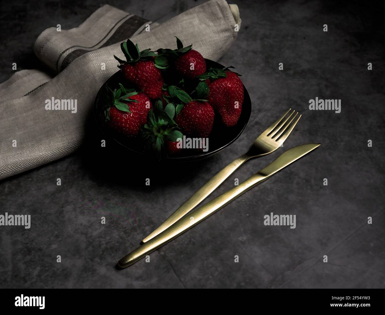Fresh strawberries in a bowl with golden cutlery and cloth napkin. Dark food style. Stock Photo