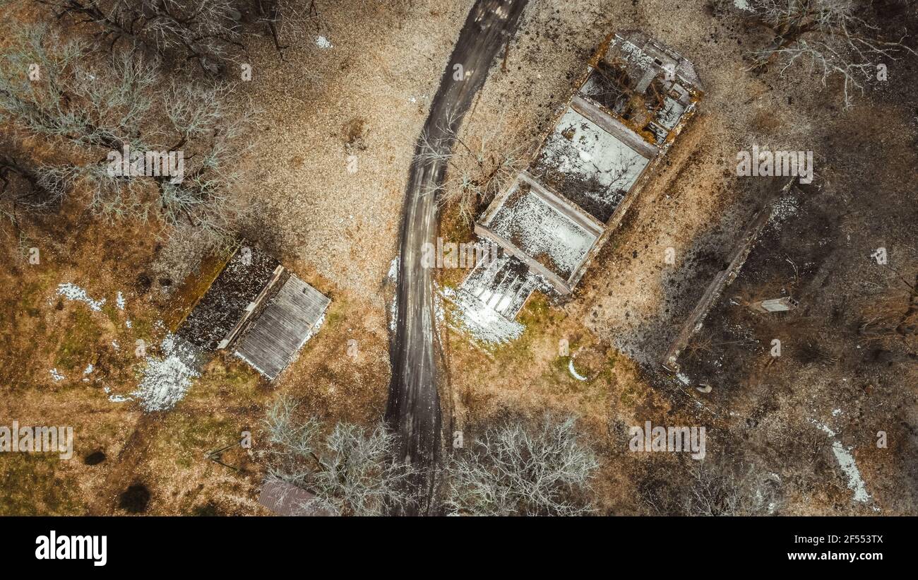 An aerial shot of a rural landscape with a dirt road across a meadow with an abandoned building with a collapsed roof and withered trees. Early spring Stock Photo