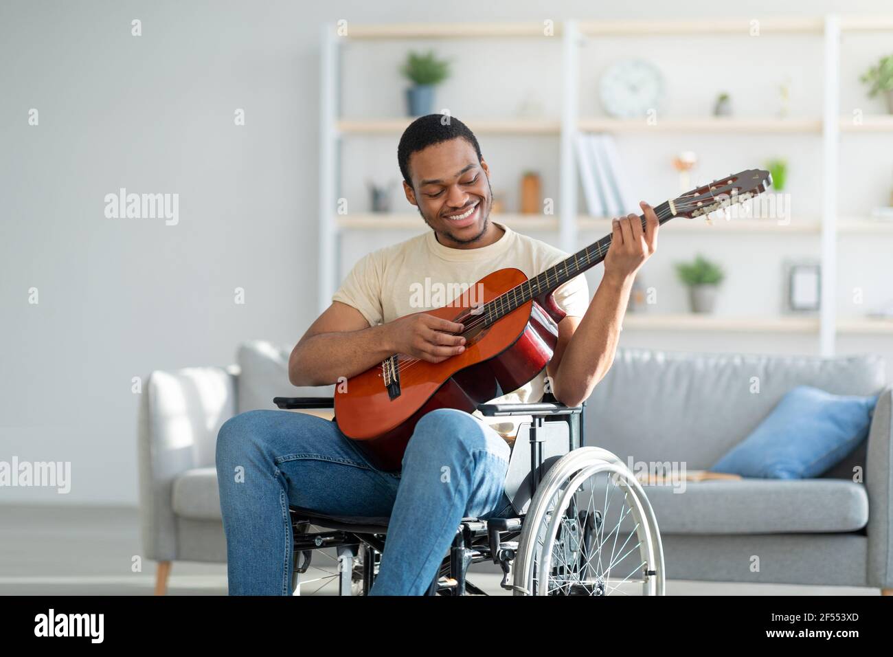 Handicapped black guy in wheelchair playing guitar and smiling at living room. Stay home activities Stock Photo