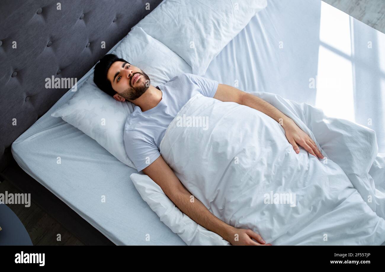 Unhappy arab man lying in bed with alarm clock, not willing to get up in  morning and suffering from headache Stock Photo - Alamy