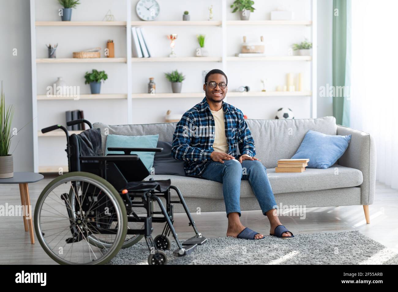 Happy disabled black guy sitting on sofa with books, empty wheelchair