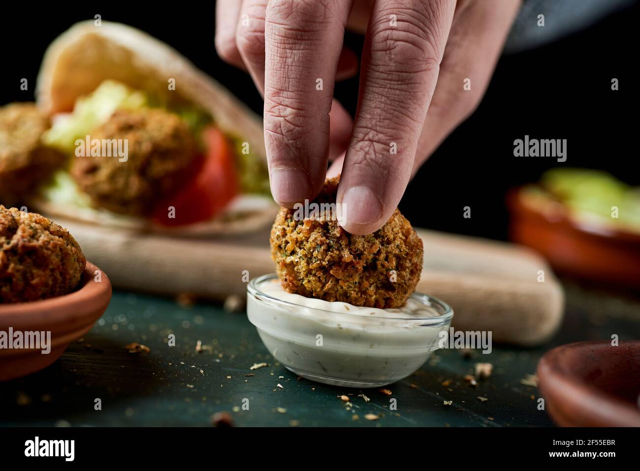 a young man dips a falafel in a bowl of yogurt sauce sitting at a rustic green wooden table Stock Photo
