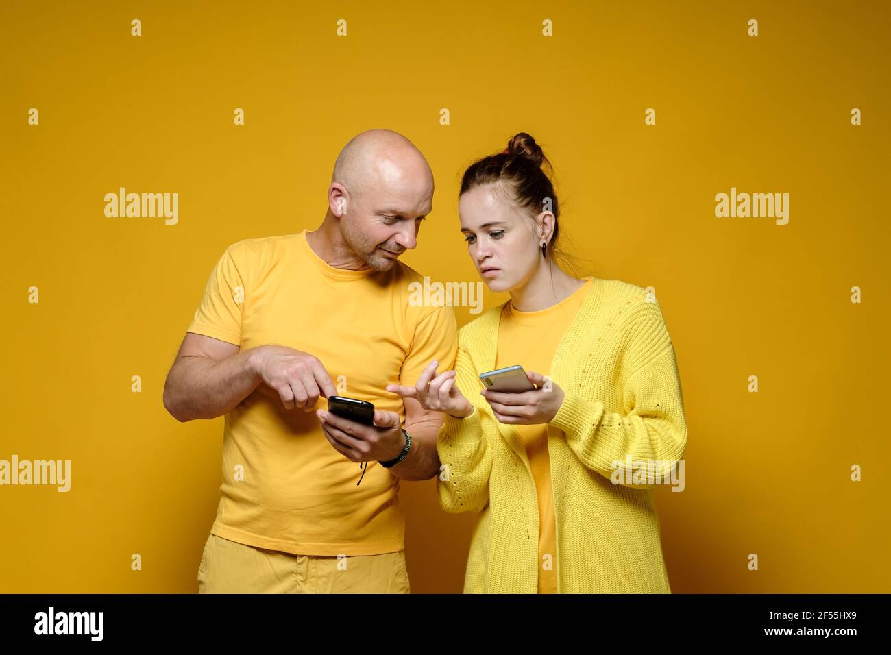 A man and a woman with a serious expression on their faces are looking at each other's smartphone screens. Yellow background. Stock Photo