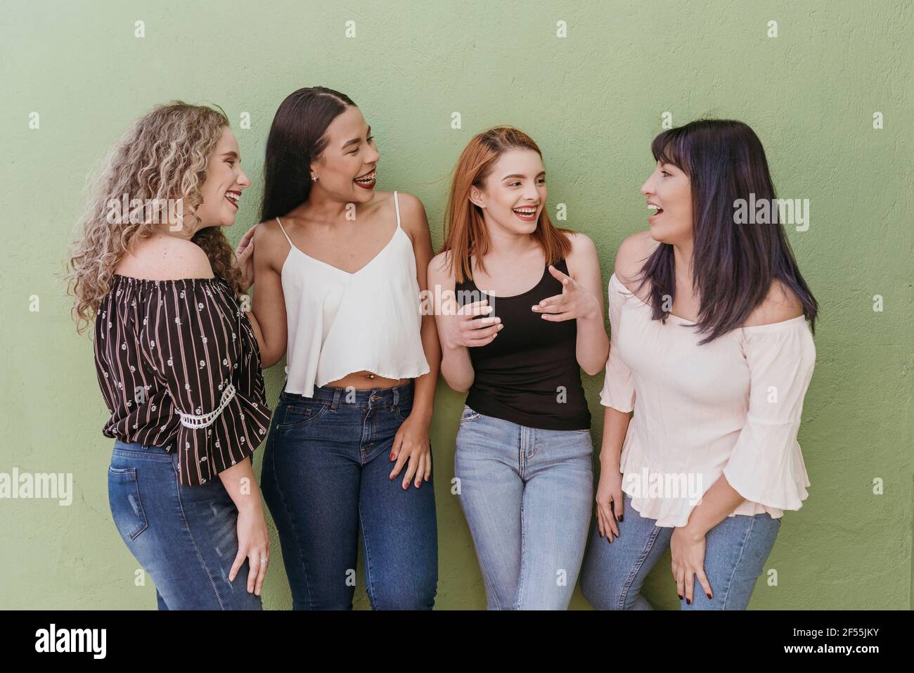 Multi ethnic female friends gossiping while standing against green wall Stock Photo