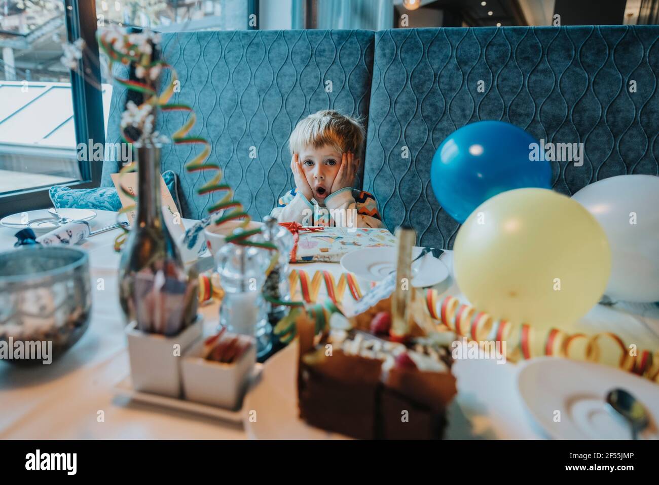 Surprised boy with hand on cheek sitting at table during birthday celebration in hotel Stock Photo