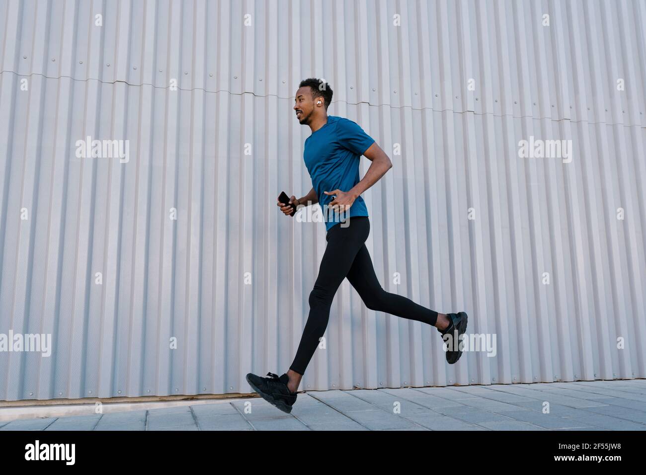 Male athlete with mobile phone running on sidewalk by wall during training Stock Photo