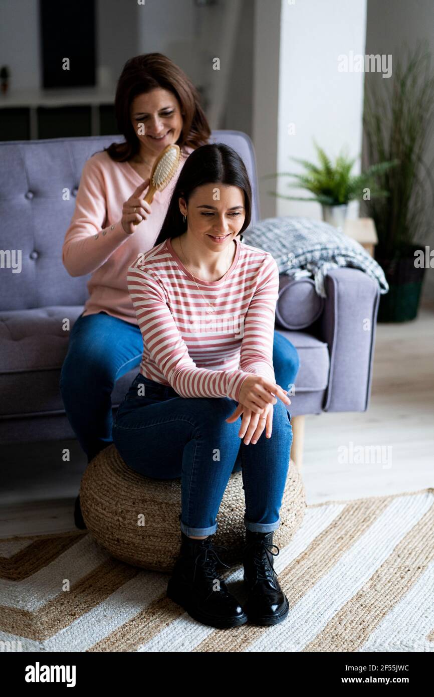 Smiling mother combing daughter's hair at sofa in living room Stock Photo