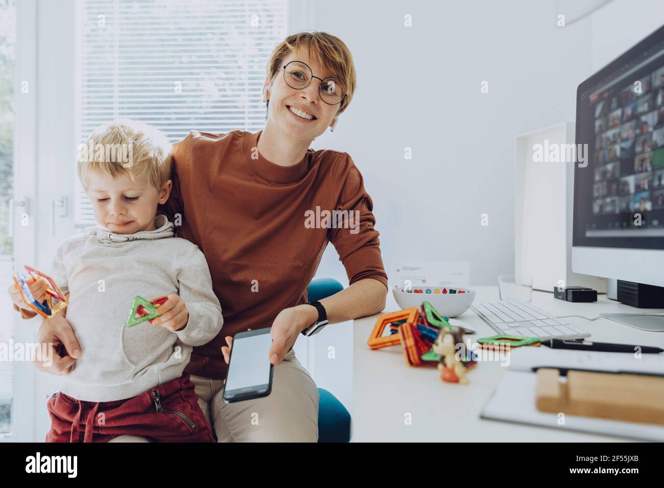 Smiling mother with son sitting on lap playing with toys in home office Stock Photo