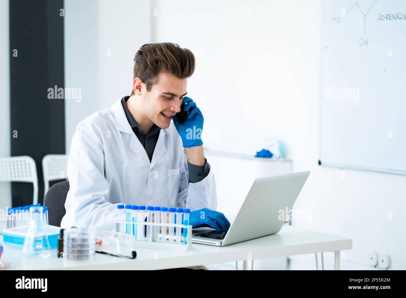 Male scientist talking on smart phone while using laptop at desk in laboratory Stock Photo