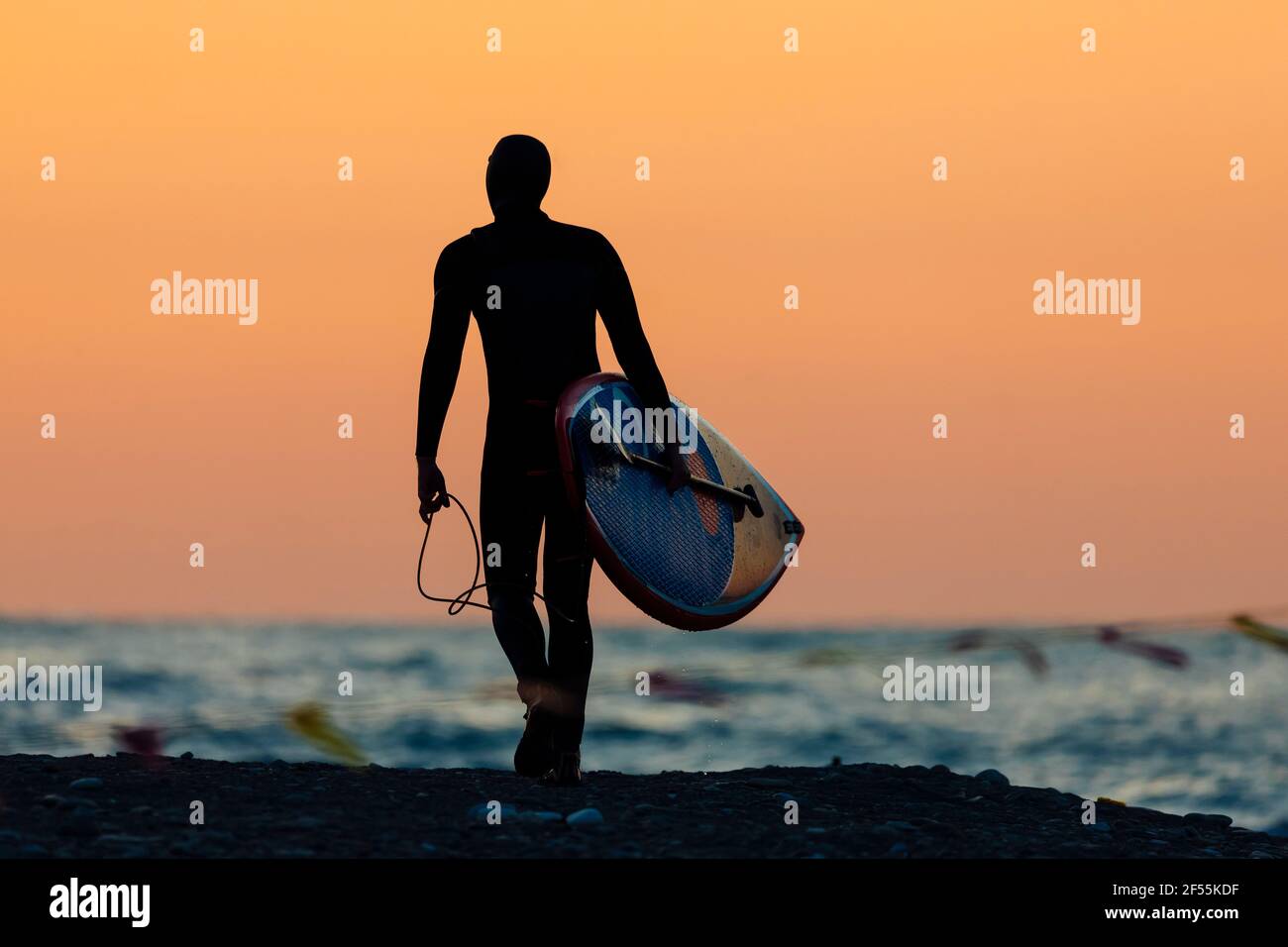 Silhouette of male surfer walking against moody sky at dusk with surfboard in hand Stock Photo