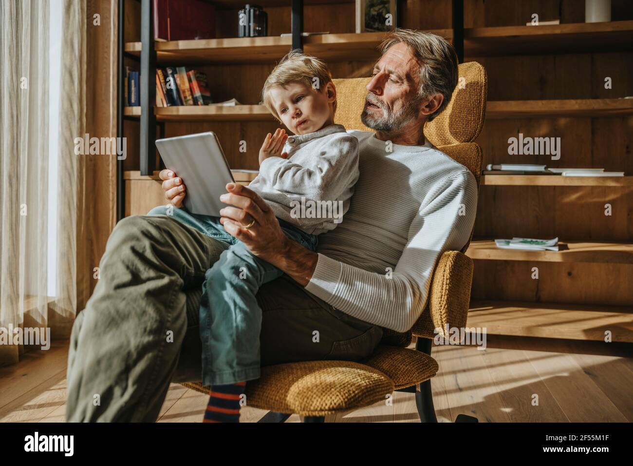 Boy E-learning through digital tablet while sitting on father's lap on chair at home Stock Photo