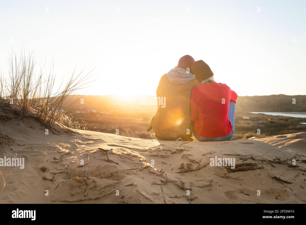 Woman leaning on man shoulder while sitting on sand dune during sunset Stock Photo