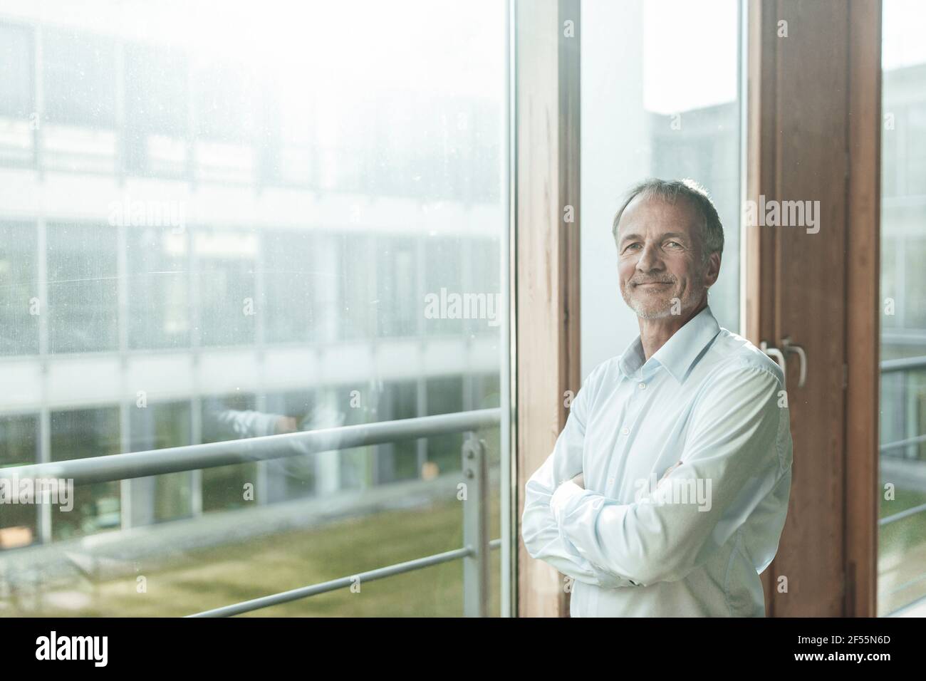 Smiling male entrepreneur with arms crossed standing at glass window in ...