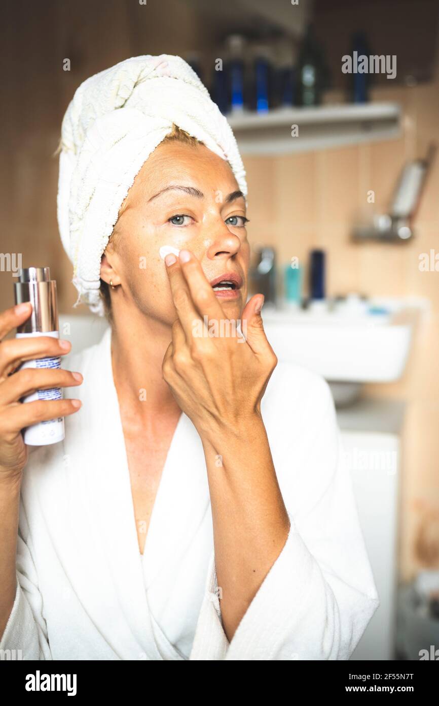 Mature woman in bathrobe moisturizing face in bathroom Stock Photo